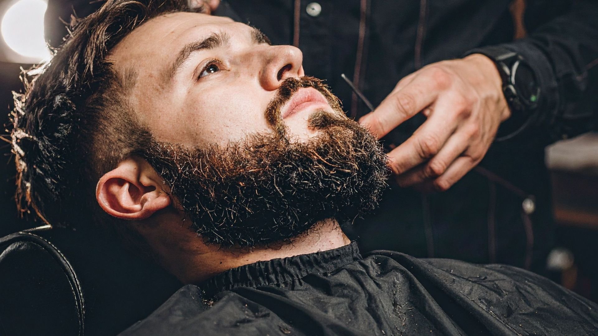Barber trimming and styling thick beard of client in barbershop