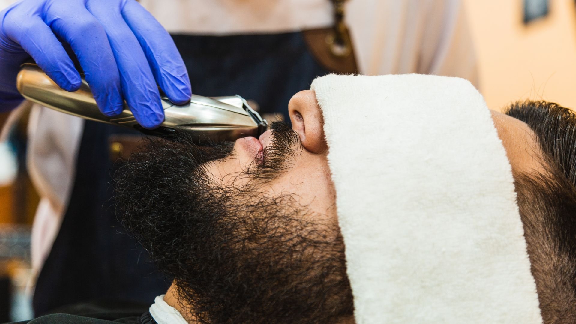 Barber trimming facial hair with electric razor, wearing blue gloves