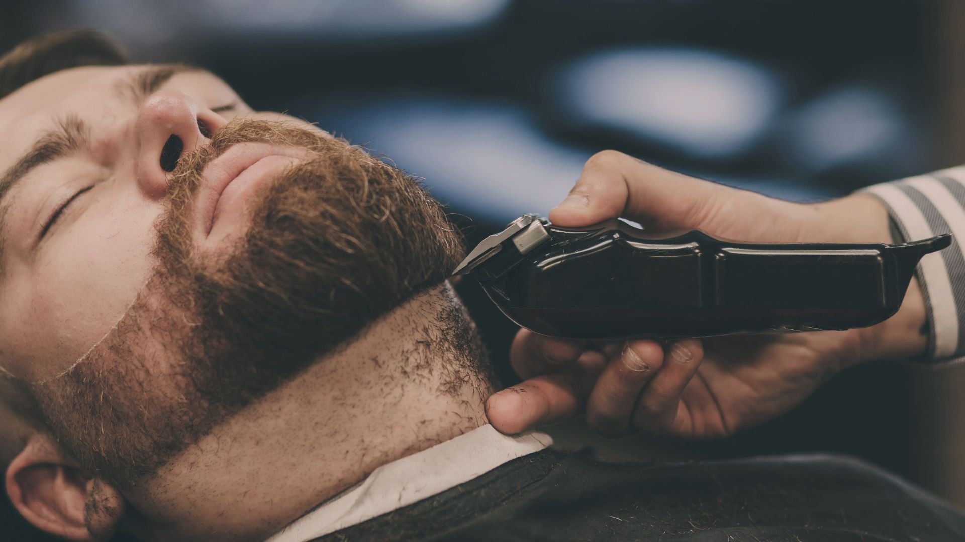 Bearded man getting beard trimmed with electric clipper at barbershop